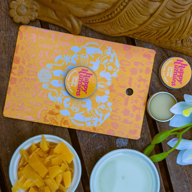 An orange and white decorative tin of Happy Mammoth body butter is displayed on a wooden table alongside bowls of ingredients (mango chunks and white balm) and a white orchid flower. A second open tin of body butter and its matching lid are also visible.