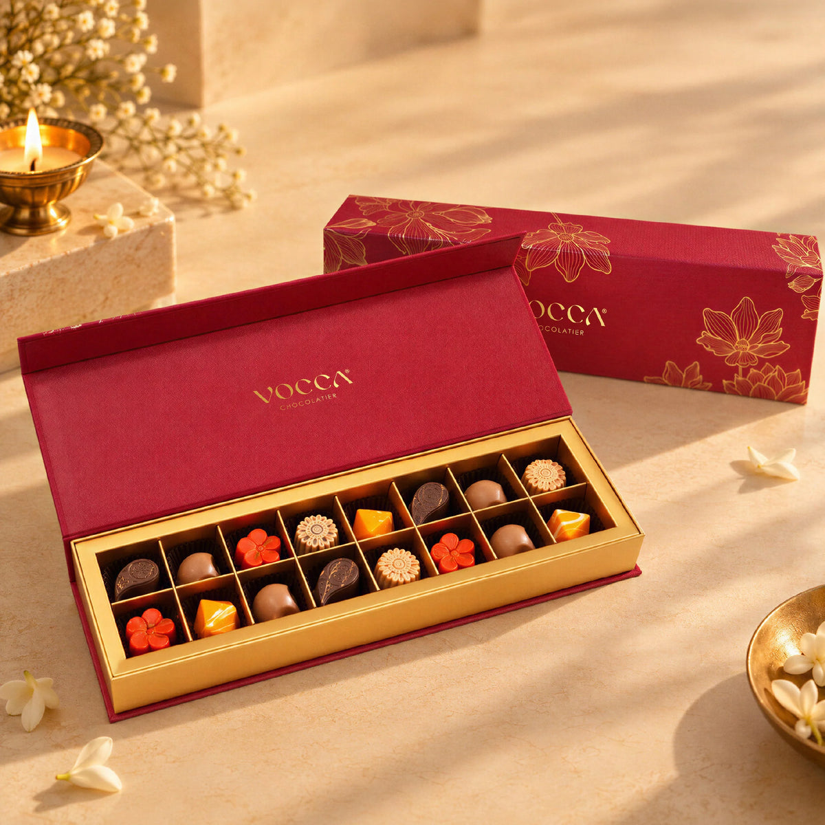 An open box of VOCCA gourmet chocolates displayed next to its matching red and gold packaging box on a warm beige surface with soft decorative props.