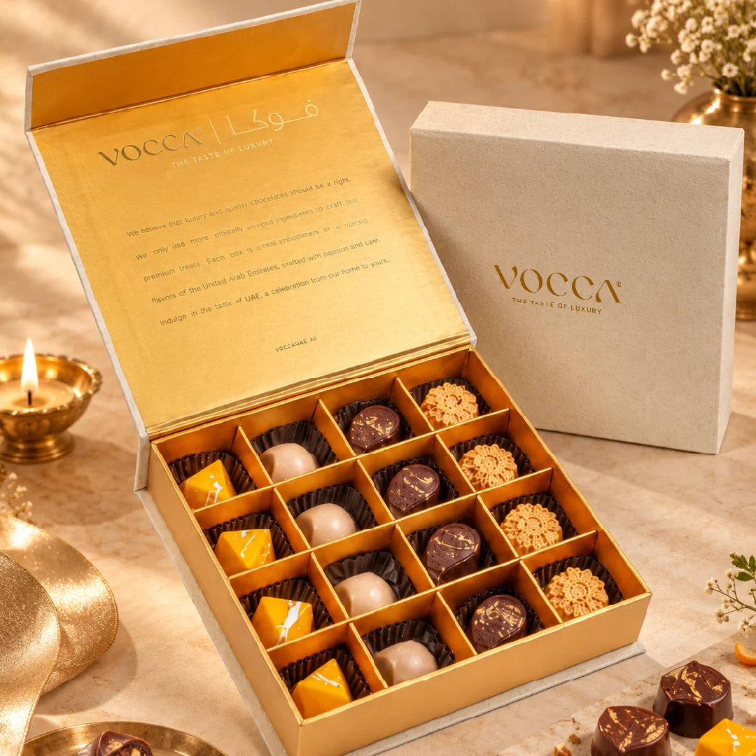 An open VOCCA chocolate gift box filled with assorted chocolates, shown beside a closed matching VOCCA box on a marble surface with candles and decorative elements.